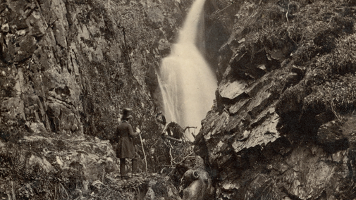 A black and white vintage photograph of a waterfall cascading through a gully with a man in a tall black hat standing looking at it